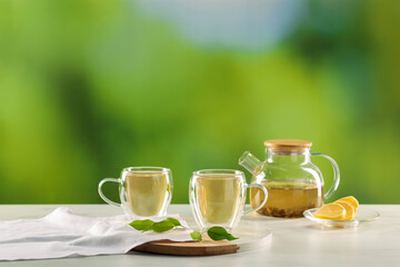 Tasty green tea in glass cups, teapot, lemon and leaves on white marble table outdoors