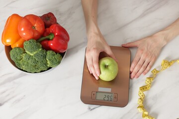 Woman weighting apple on kitchen scale at white marble table, top view