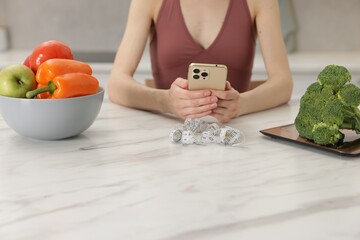 Woman using smartphone at white marble table with vegetables and measuring tape indoors, closeup