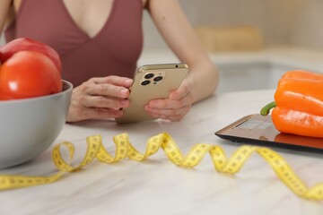 Woman using smartphone at white marble table with vegetables, kitchen scale and measuring tape...