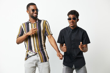 Two cheerful Black men dance joyfully together, wearing stylish sunglasses and colorful shirts, against a bright white background, showcasing friendship and celebration.