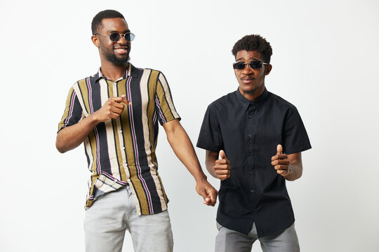 Two cheerful Black men dance joyfully together, wearing stylish sunglasses and colorful shirts, against a bright white background, showcasing friendship and celebration.