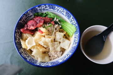 A colorful bowl of dry noodles topped with vegetables, wonton crisps, and barbecued meat, served...