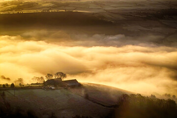 Golden mist envelops a valley, with hilltops and buildings rising above the ethereal clouds in a...