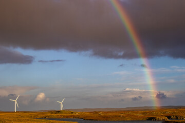 Vibrant rainbow arcs across a sky with dark clouds and blue patches, illuminating a landscape with...
