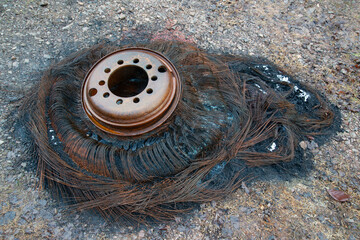 Rusted metal hub of a wheel rests on the charred, tangled remains of a burnt tire, revealing melted...