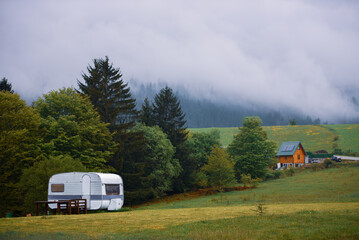 A camper van parked on a colorful grassy hill, with a small cabin and dense fog rising from the...