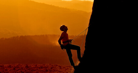 A climber in a red shirt and helmet scales a steep rock face at sunset. The silhouette of the climber contrasts against the warm, golden sky.