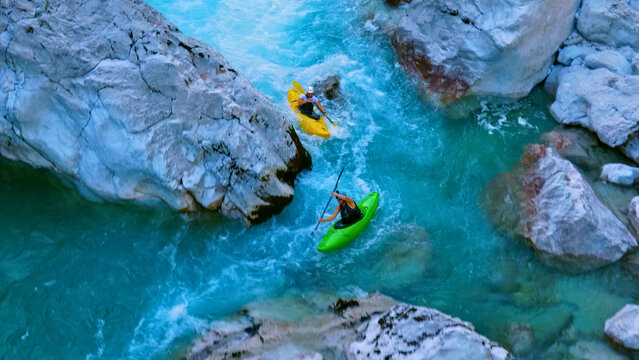 A thrilling aerial view showcases two kayakers navigating a narrow, fast-flowing river between large rocks. One kayaker, wearing a white helmet, paddles a yellow kayak, while the other, in a black.