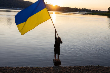 child holds large yellow-blue flag and waves it standing in Dnieper river at sunset. Proud to be...