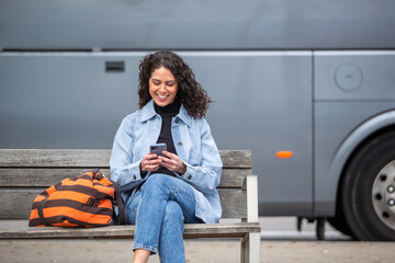 cheerful young woman sits on a wooden bench beside a large grey bus, smiling while looking at her...
