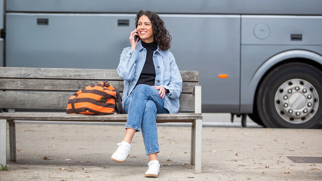 Full body young woman sitting on a bench at a bus stop, cheerfully talking on mobile phone