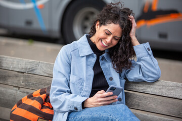 Smiling woman sitting on a bench, laughing while looking at her phone, wearing jacket and jeans,...