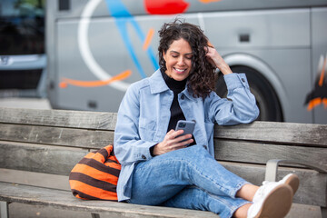Smiling woman sitting on a bench using her smartphone, with travel bag, relaxed near a parked bus.