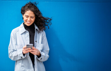 Young woman smiling while using phone, standing against a vibrant blue wall with the wind blowing...