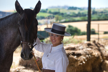A young, beautiful woman dressed in riding clothes and a traditional Spanish hat is standing next to her grey horse in the barn. The woman is in love with her horse and touches it.