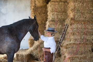 A young, beautiful woman dressed in riding clothes and a traditional Spanish hat is standing next to her grey horse in the barn. The woman is in love with her horse and touches it.