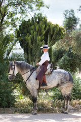 A young, attractive Spanish woman dressed in riding attire and wearing a traditional Spanish hat, riding a horse in the countryside on a sunny day. In the background, green vegetation.
