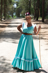 A beautiful young Spanish woman dressed in a traditional green flamenco dress and with a flower in her hair walks through the pine forests of Andalusia in the pilgrimage, leaning on a stick.
