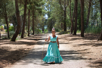 A beautiful young Spanish woman dressed in a traditional green flamenco dress and with a flower in her hair walks through the pine forests of Andalusia in the pilgrimage, leaning on a stick.