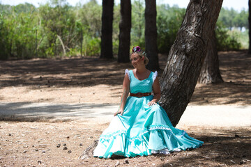 A beautiful young Spanish woman dressed in a traditional green flamenco dress with a flower in her hair rests seated on a pine tree trunk at a typical Andalusian pilgrimage.