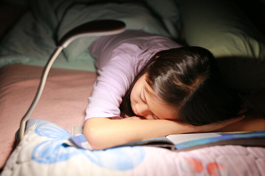Portrait of Asian girl kid sleeping while reading a book under lamp light on the bed at night. Close up shot and soft focus.