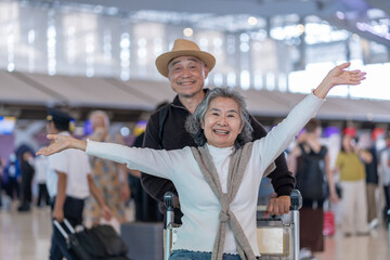 happy asian senior woman sitting on luggage that put on trolley,raising hands up,her husband standing behind in airport,excited and ready to travel