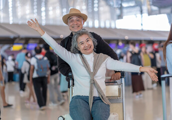 happy asian senior woman sitting on luggage that put on trolley,raising hands up,her husband standing behind in airport,excited and ready to travel
