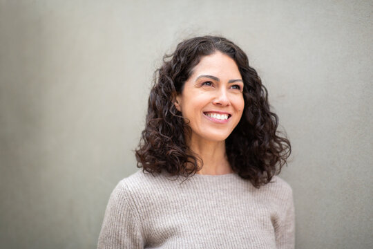 Portrait of a cheerful woman with curly hair standing against a light gray wall, smiling brightly and looking away