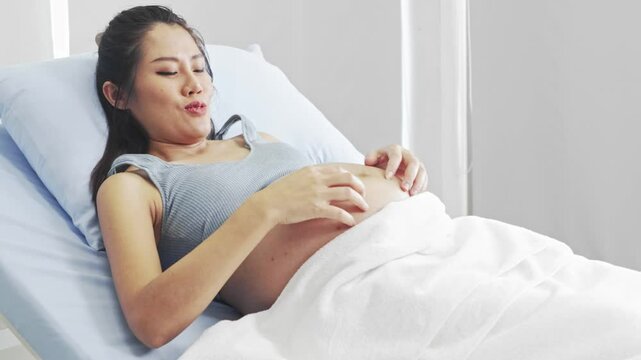 Pregnant Asian woman sits on hospital bed gently scratching and rubbing her belly with both hands relieve itching caused by skin expansion dry cracking and rashes waits doctor to check her pregnancy.
