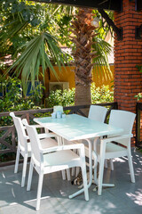Outdoor dining area with white furniture surrounded by lush greenery, palm trees. Cat under table