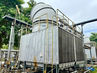 Industrial cooling tower system emitting vapor under blue sky. Heavy-duty piping, valves, and heat exchangers in a manufacturing or refinery plant exterior.