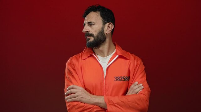 Hispanic man with a beard wearing an orange prison jumpsuit stands against a bold red background, exuding a serious demeanor.