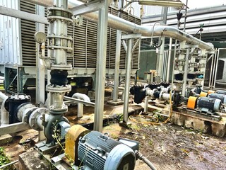Industrial cooling tower system emitting vapor under blue sky. Heavy-duty piping, valves, and heat exchangers in a manufacturing or refinery plant exterior. Water pumps 