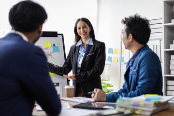 Asian businesswoman presenting business plan on whiteboard during a meeting. Team of businesspeople is listening to their coworker explaining a business plan during a work meeting in office.