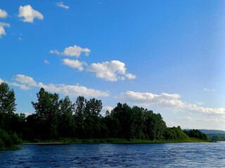 Calm river under a bright blue sky with fluffy clouds and lush trees along the shoreline during the...