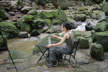 Relaxing by the stream, a woman enjoys nature while sitting on a foldable chair among mossy rocks...