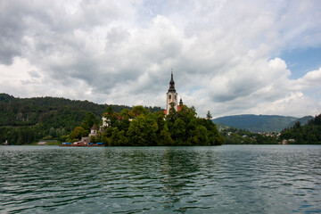 Naklejka premium The iconic Church of the Assumption of Mary, with its distinctive tall belfry and white and red facade, stands majestically amidst lush green trees on Bled Island
