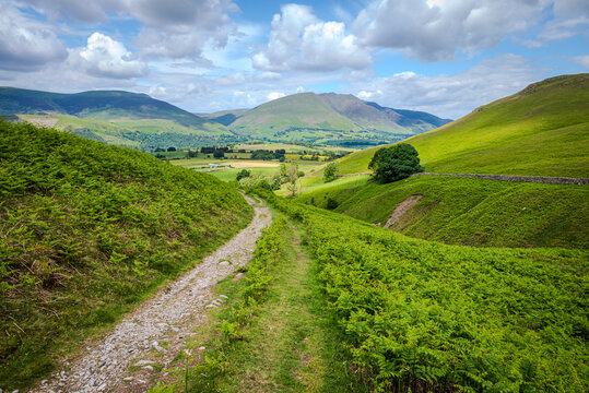 View of a hiking path meandering through a lush green valley nestled between curving fells, with a mountain range rising in the distance in the Lake District
