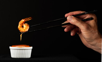 Single shiny grilled shrimp held by chopsticks with orange sauce dripping over white ramekin...