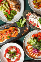Assorted restaurant-style dishes including grilled fish, chicken with vegetables, creamy tomato salad, and fried patties with cured meat, all artfully presented for food photography.