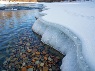 "A late-winter scene where a thin sheet of ice clings to the edge of a wide river bend, clear water exposing colorful smooth pebbles below, small meltwater rivulets forming silvery lines across 