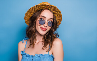 Portrait of a beautiful young woman in a blue dress, straw hat, and sunglasses, posing against a pastel background. She is looking at the camera.