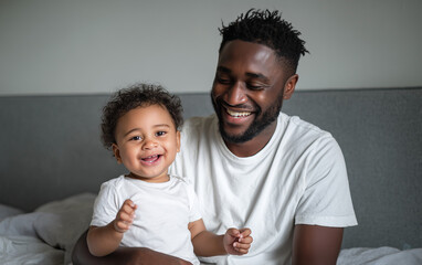 A Black father wearing a white t-shirt playing with his baby son in the bedroom, both smiling and happy. The image was captured using studio lighting,