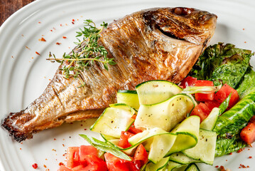 Grilled fish with thyme, zucchini ribbons, tomatoes, and lettuce on a white plate. Healthy gourmet meal with fresh vegetables, perfect for clean eating and food styling.