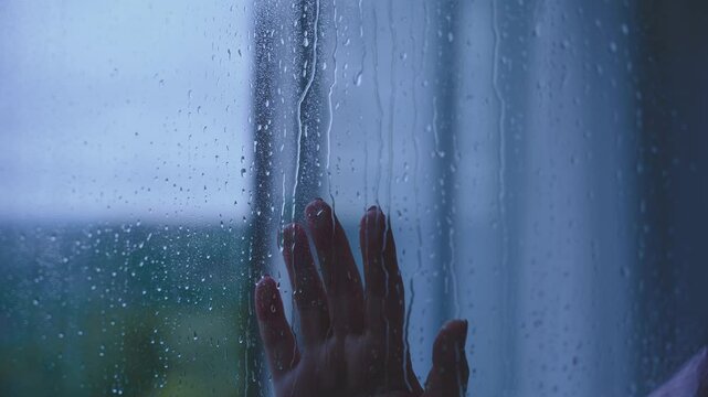 Close-up hand fingers touching wet window rain drops falling water drip flow down glass. concept unhappy, mood depression sadness emotion sad unrecognizable person body part. lonely people. dark room.