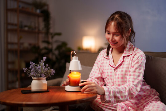 Asian young adult woman in comfortable pajamas enjoys a tranquil evening creating a serene ambiance with a glowing candle warmer for personal well being and relaxation 