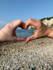 Hands forming a heart shape against a scenic seaside backdrop with cliffs, pebbles, and calm...