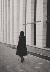 A woman in a long coat and high heels walks alone by a modern building with tall columns, captured in a black and white photo © Alissa Fuhrmann