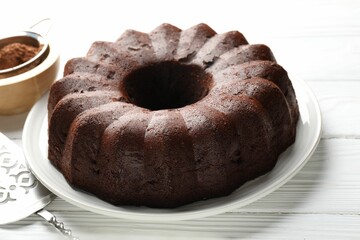 Tasty chocolate bundt cake, cocoa powder and server on white wooden table, closeup